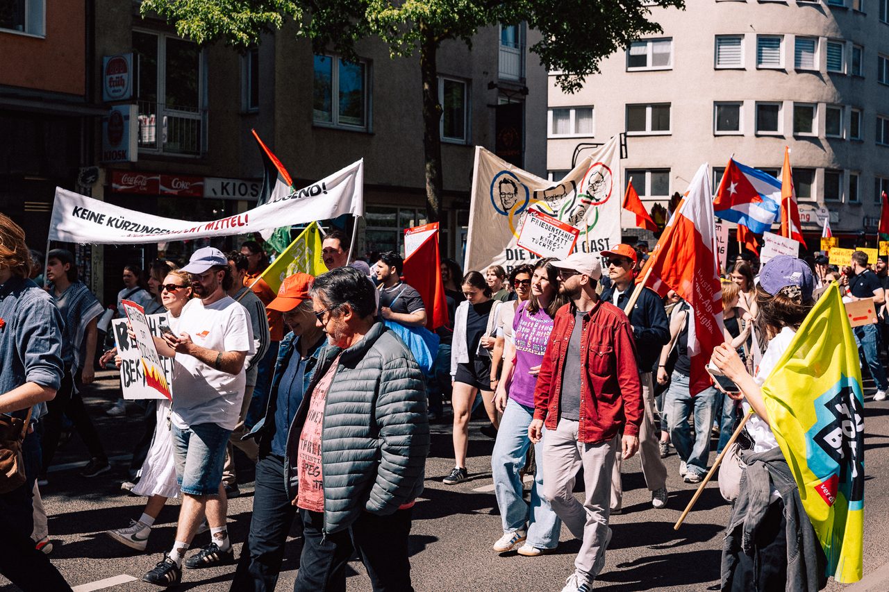 Demonstrierende ziehen mit Fahnen und Schildern durch eine sonnige Stadtstraße
