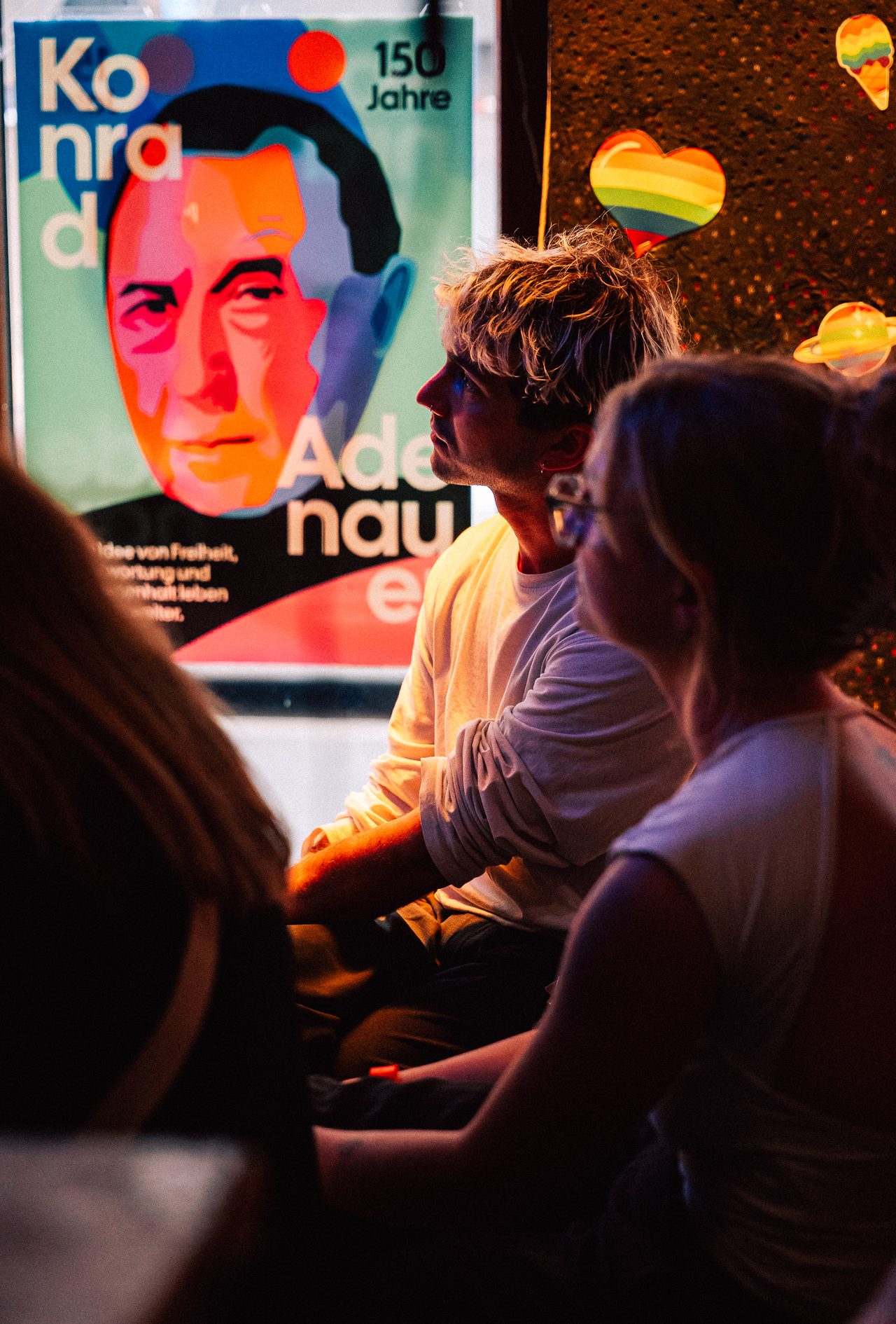 Sitzende Gäste hören vor einem bunten Adenauer-Plakat aufmerksam zu