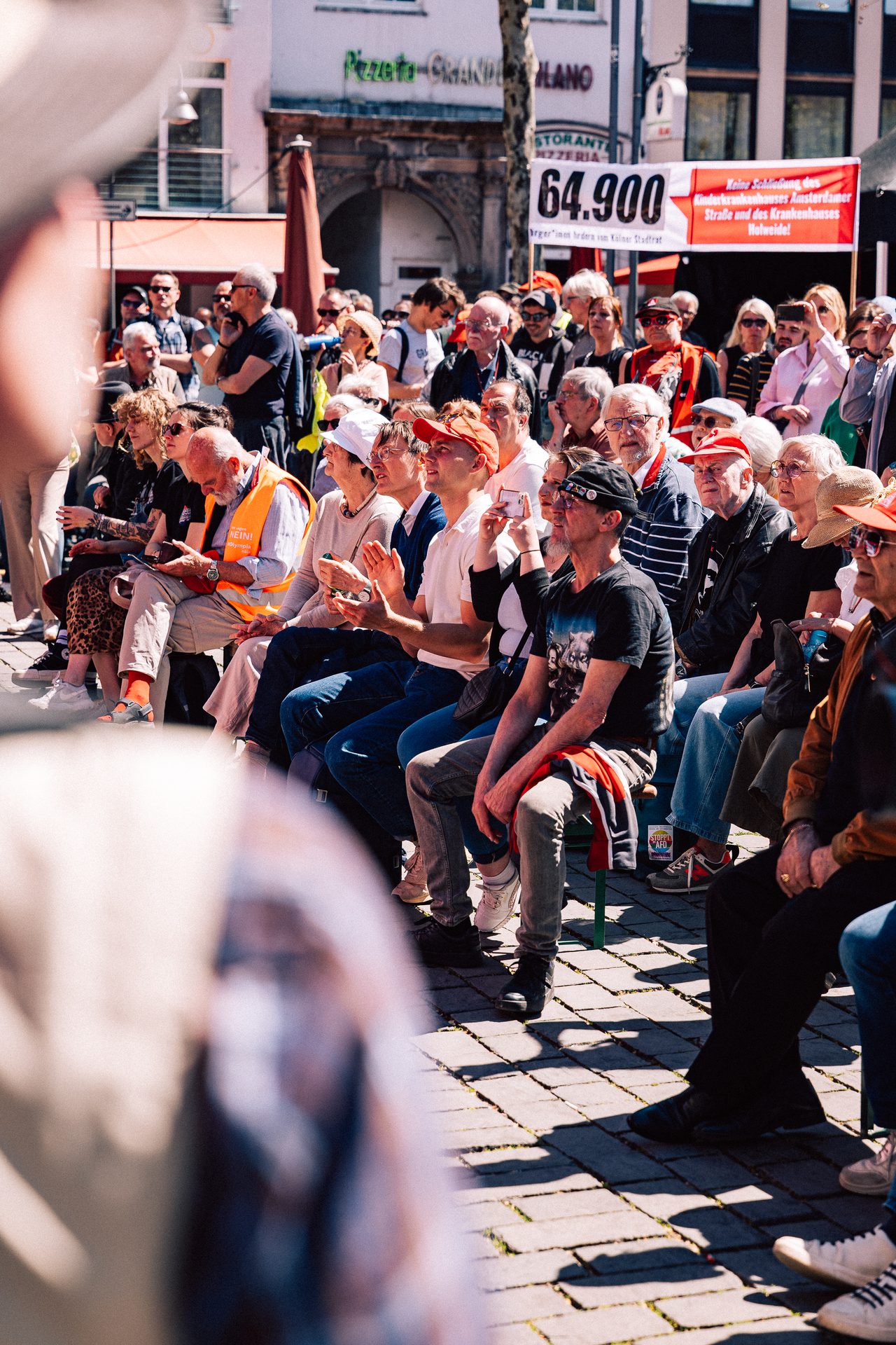 Sitzendes Publikum verfolgt eine Kundgebung auf einem sonnigen Stadtplatz